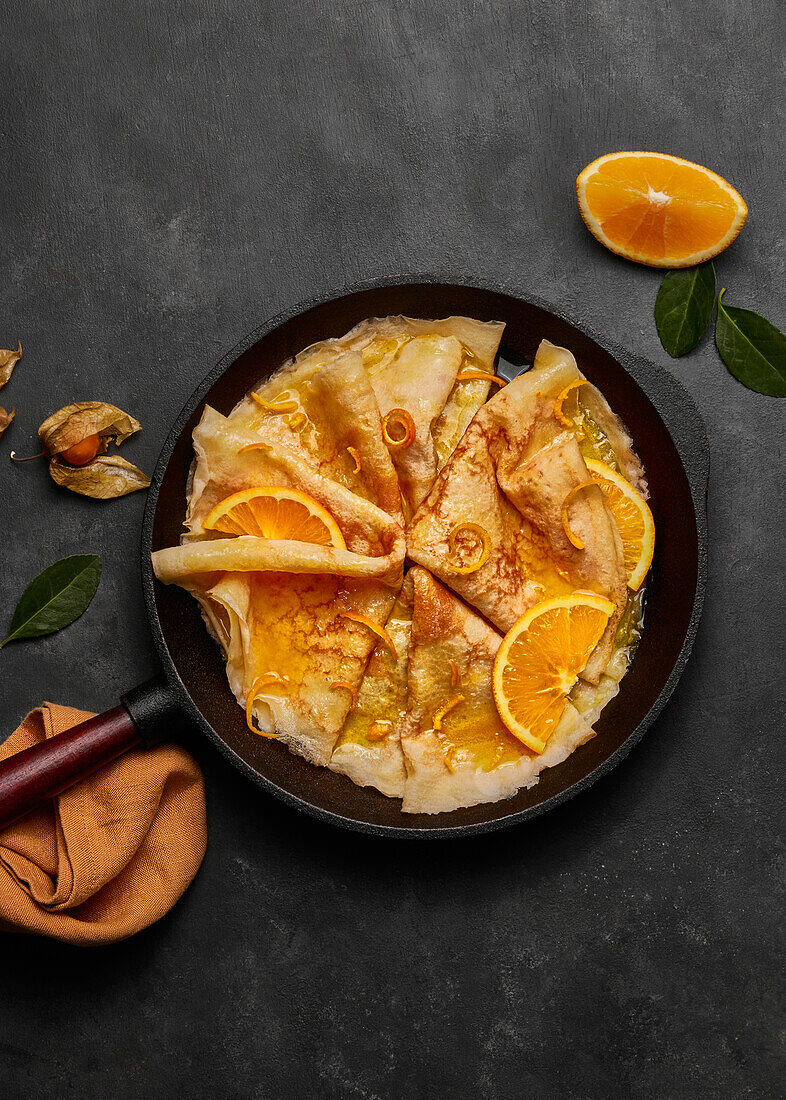 From above homemade crepes suzette with orange liqueur and orange slices served on plate with knife and fork on concrete surface