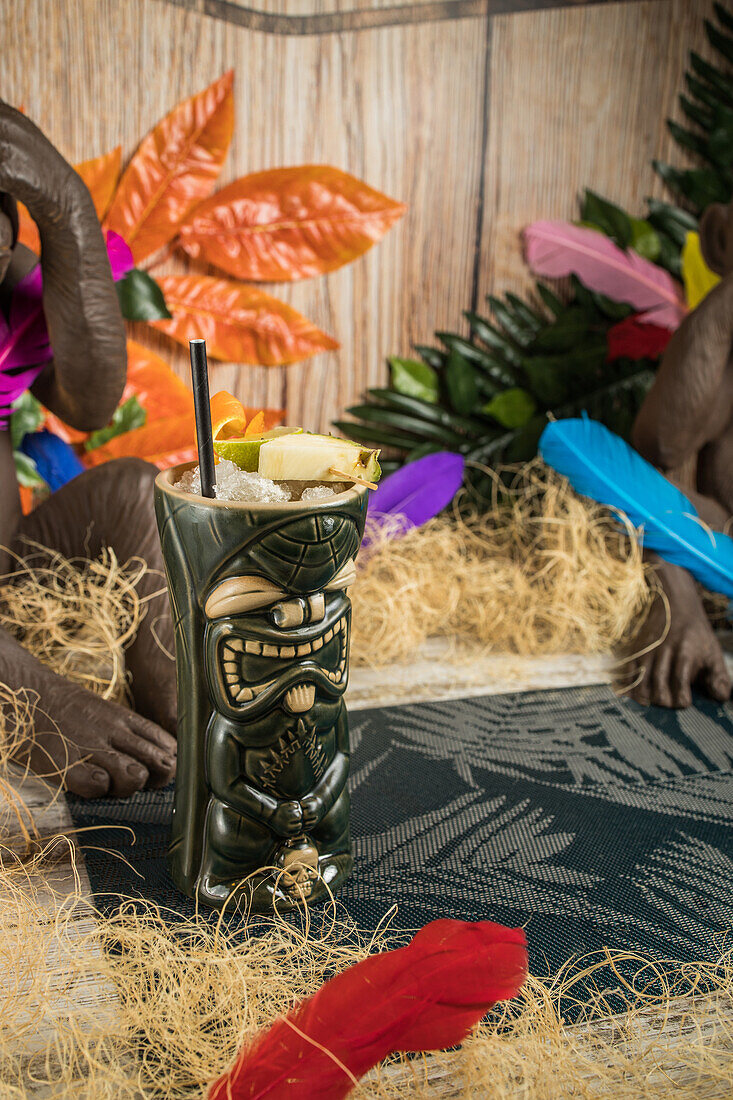 From above of large sculptural tiki cup filled with booze decorated with straw and fruits placed on green rug against dry grass