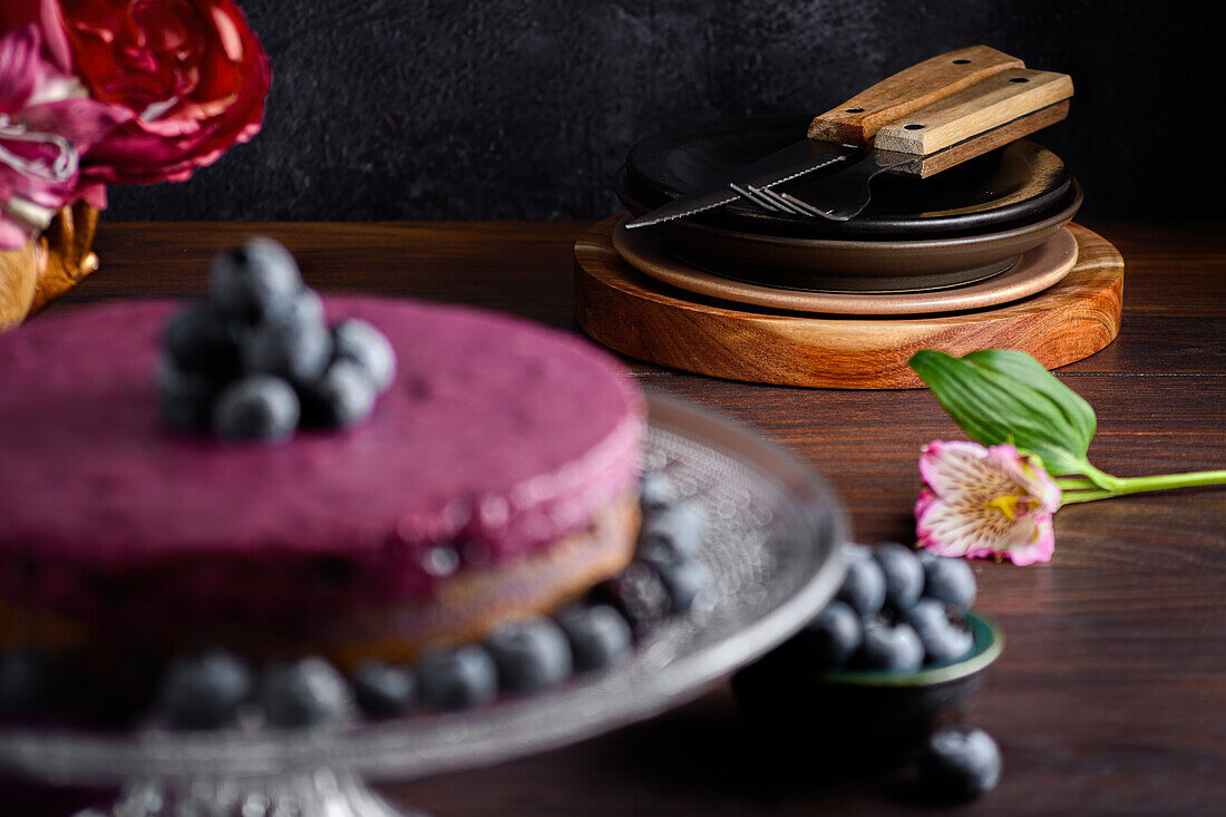 Delicious blueberry mousse cake with purple cream decorated with fresh berries served on glass stand on dark table with flowers