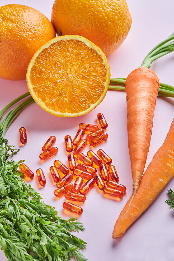 Von oben Komposition von verstreuten Vitaminpillen auf rosa Tisch neben reifen Karotten und saftigen Orangen angeordnet