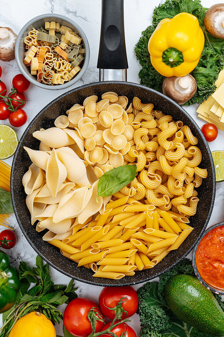 From above of frying pan with various uncooked pasta surrounded with assorted raw vegetables on marble table in light kitchen