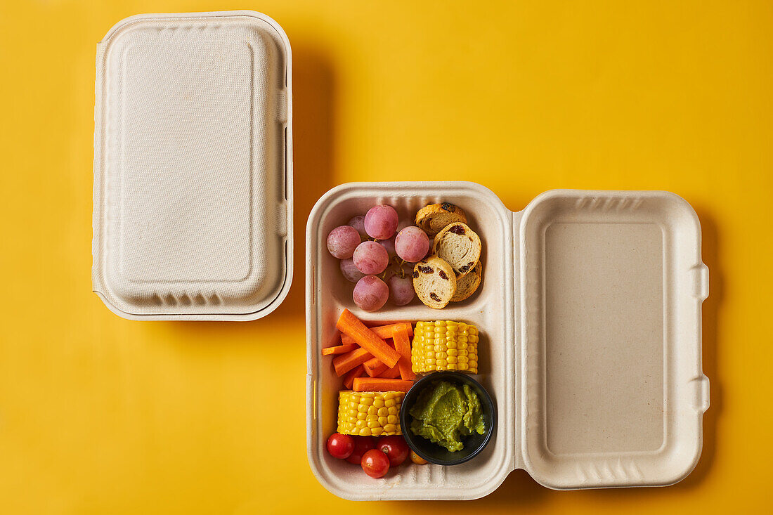 From above of lunch boxes with healthy food including crackers carrot sticks grapes cherry tomatoes with broccoli walnut and tangerine on yellow background