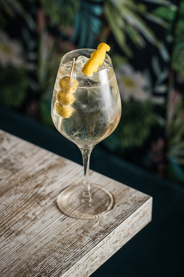 Blick von oben auf ein Kristallweinglas mit einem Martini-Cocktail, serviert mit Zitronenschale und Oliven am Rand eines Holztisches