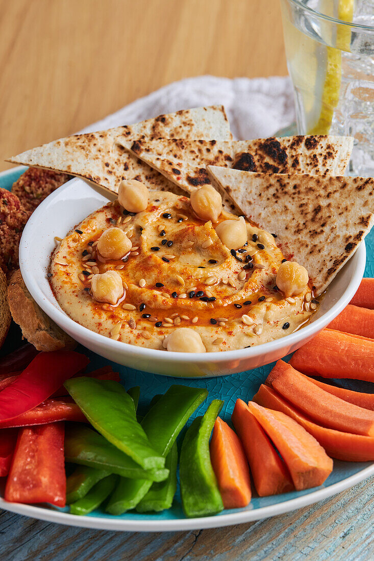 From above of yummy hummus with tortilla chips served on plate with croutons and assorted vegetables on rustic cutting board near glass of cold lemonade on wooden table with napkin