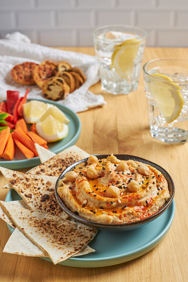 From above of delicious hummus and tortilla chips served on plate near assorted vegetables and croutons and glasses of cold lemonade on wooden table