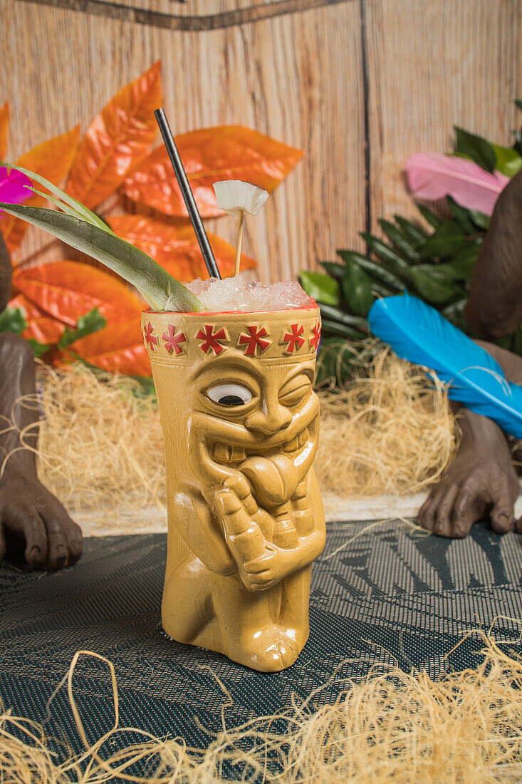 Polynesischer Tiki-Becher mit kaltem alkoholischem Getränk, dekoriert mit Stroh und grünen Blättern vor bunten Blättern und trockenem Gras