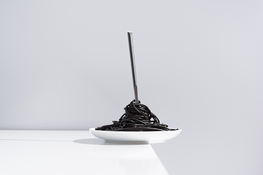 Stainless fork in full bowl of black spaghetti with cuttlefish ink on white table in studio on gray background