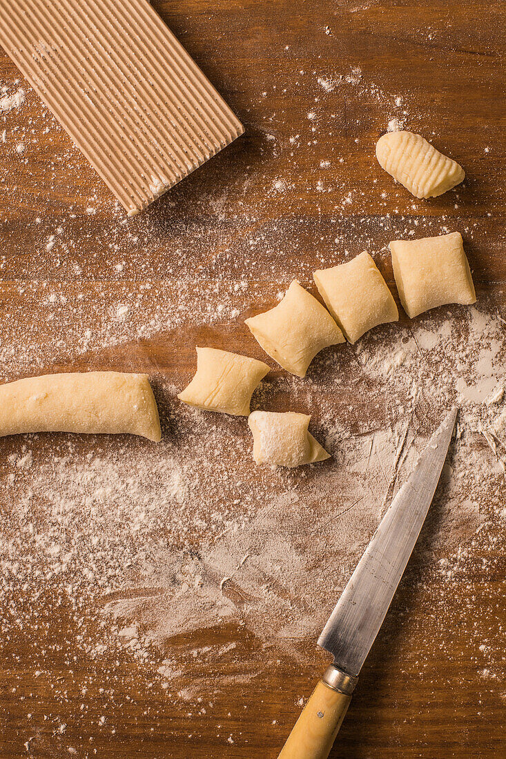 Draufsicht auf weiche, rohe Teigstücke, die bei der Zubereitung von Gnocchi in der Küche auf einem mit Mehl bestäubten Holztisch neben einem Schneidebrett und einem Messer liegen