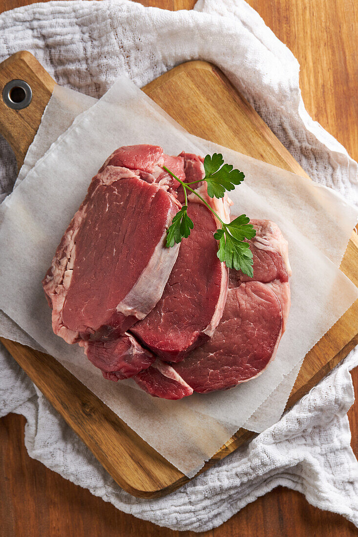 Top view slices of fresh uncooked beef fillet with green parsley placed on wooden chopping board with parchment paper in kitchen