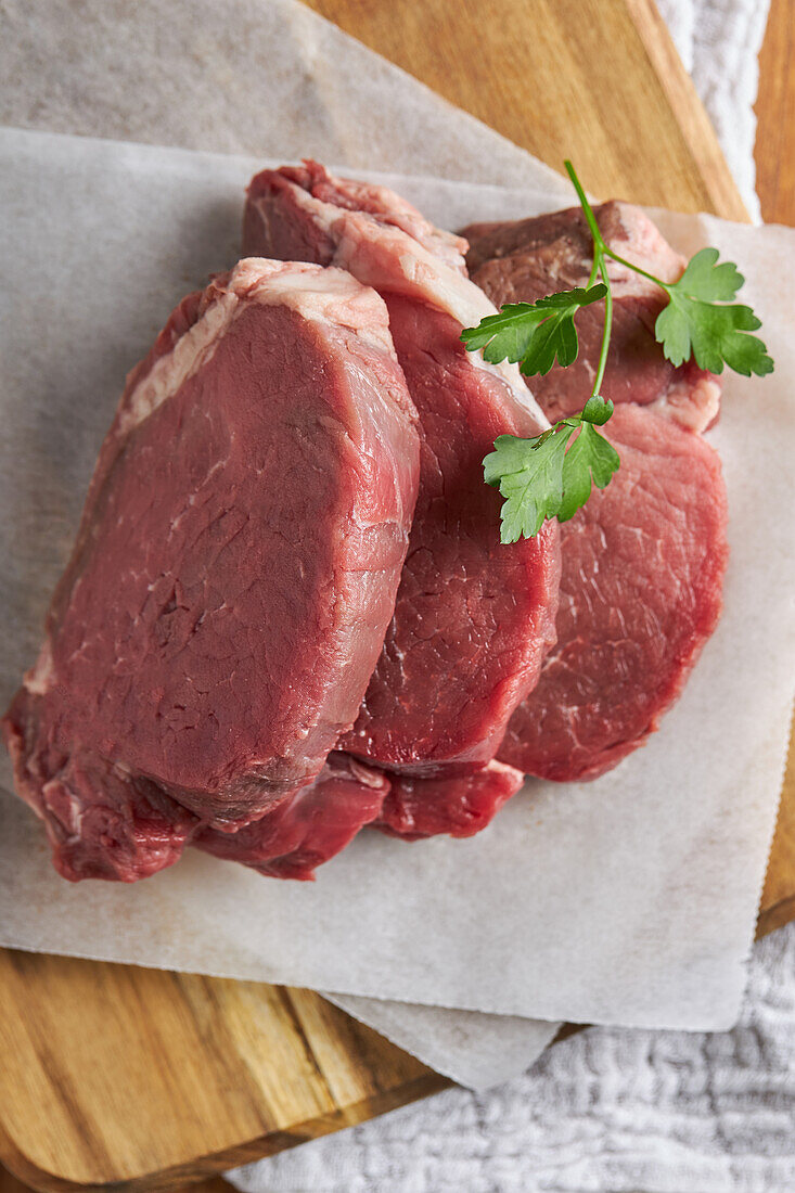 Top view slices of fresh uncooked beef fillet with green parsley placed on wooden chopping board with parchment paper in kitchen