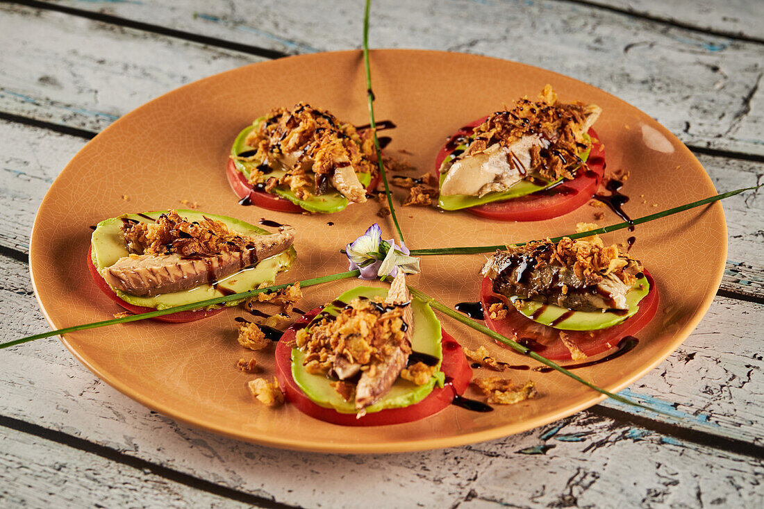 From above crispy fried onion and sliced of mackerel placed on slices of tomato and avocado near greens and flowers on plate in restaurant