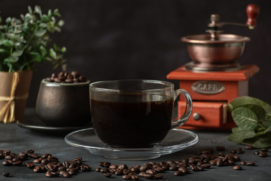 Ein Glas frisch gebrühter aromatischer Kaffee auf einer Untertasse steht auf einem Tisch mit verstreuten Bohnen vor einer manuellen Kaffeemühle