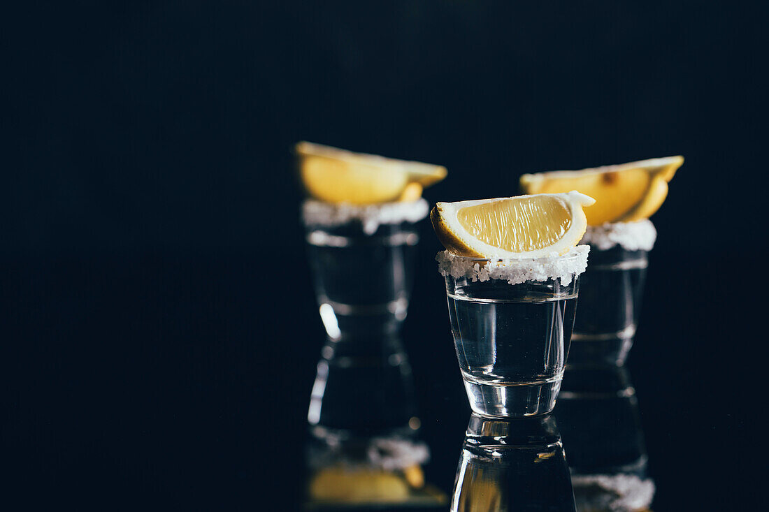 Tequila-Shots mit Salz und Zitrone auf spiegelnder Oberfläche vor dunklem Hintergrund