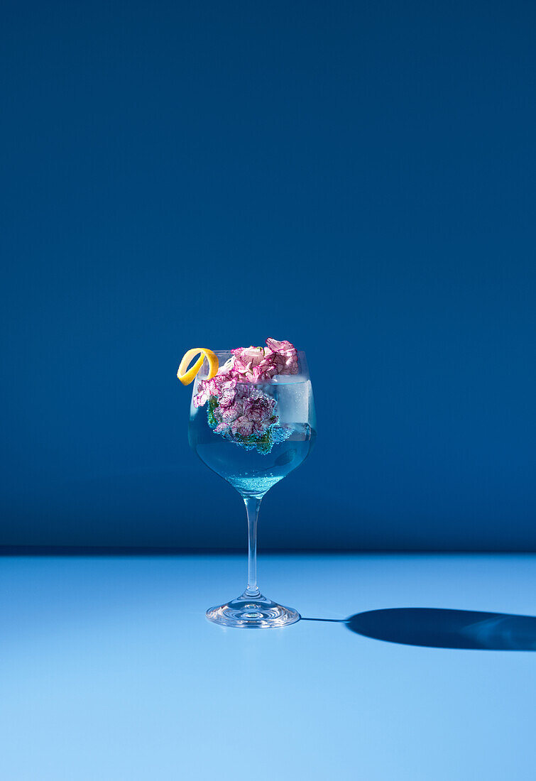 Transparentes Glas mit frischem Cocktail, Minzblättern und Blumen vor blauem Hintergrund auf dem Tisch