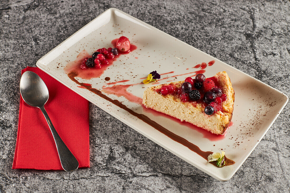 High angle of piece of delicious cheesecake with red berries and jam served on rectangular plate near glasses of water in restaurant