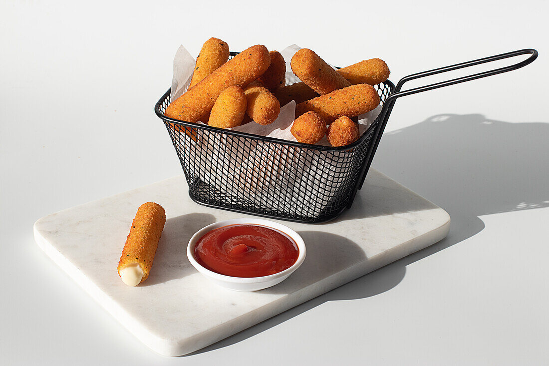 Mozzarella cheese fried sticks placed in metal fryer near ketchup sauce in marble board on white background