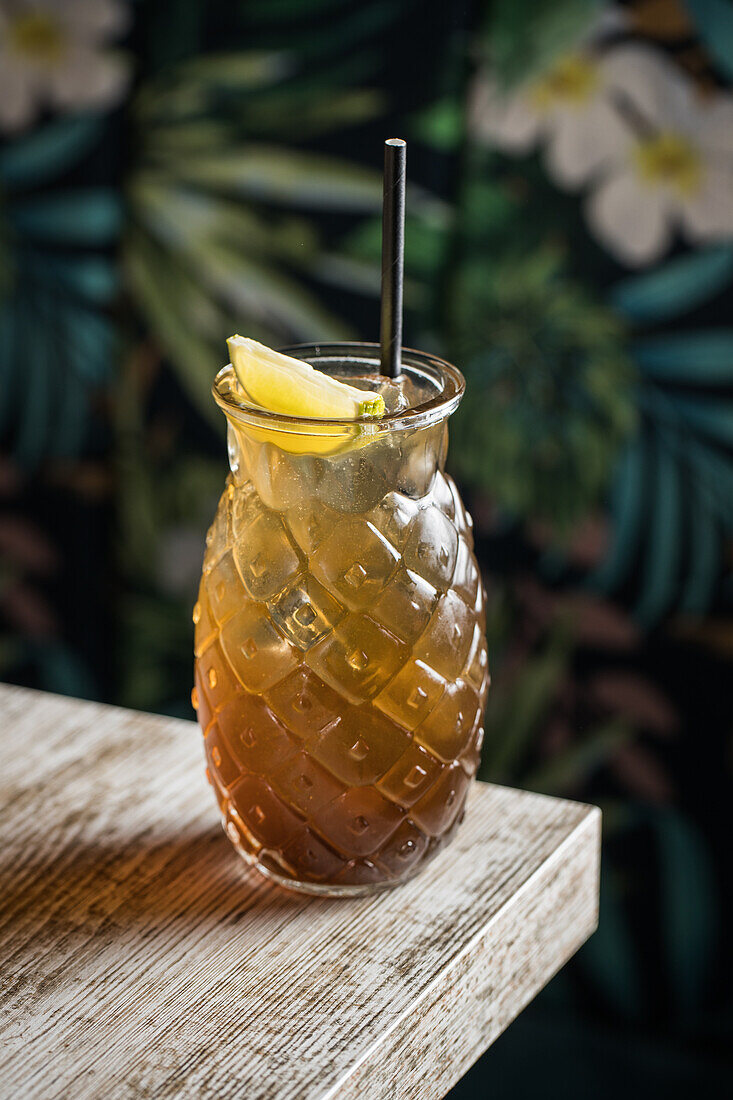 Glass tiki mug filled with alcohol beverage with straw decorated with fruit placed on edge of wooden table couch