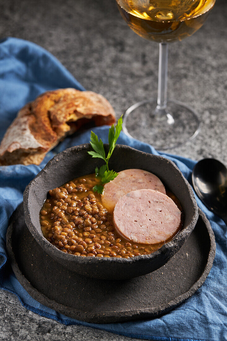 Von oben rustikale Schale mit leckerer Linsensuppe mit Petersilie und Wurstscheiben auf Marmortisch und blauer Serviette neben Brot und Wein beim Mittagessen