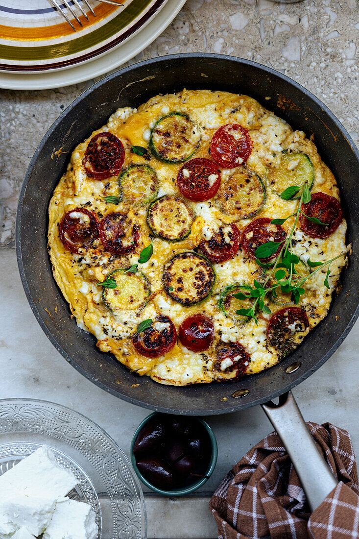 Top view of traditional Greek frittata with cherry tomatoes and feta cheese in frying pan with bread in light kitchen