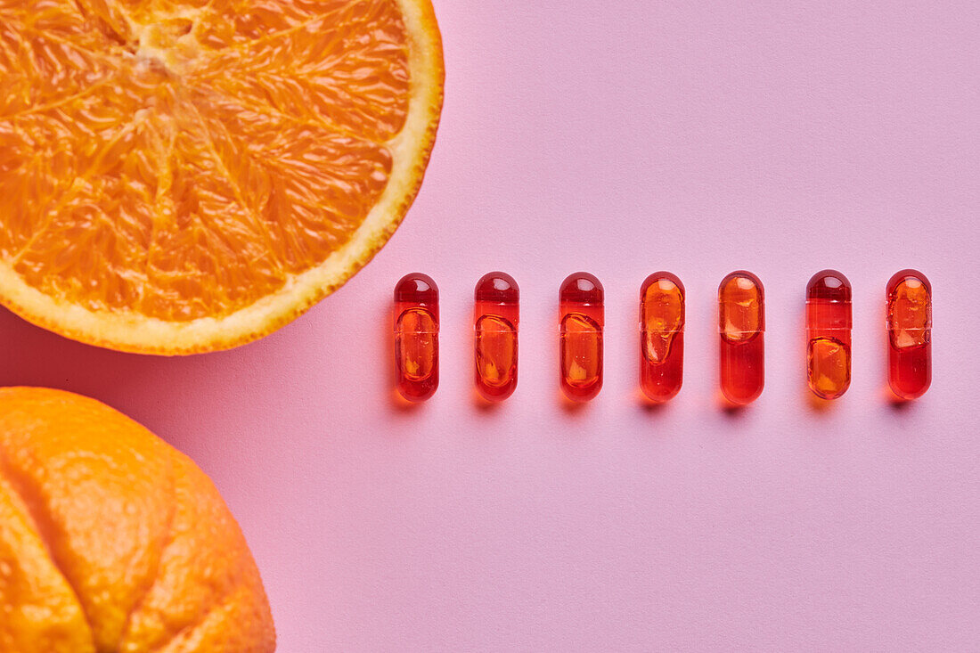 Komposition aus reifen, geschnittenen Orangen, die auf einer rosafarbenen Fläche neben einer Reihe von Pillen angeordnet sind, von oben gesehen