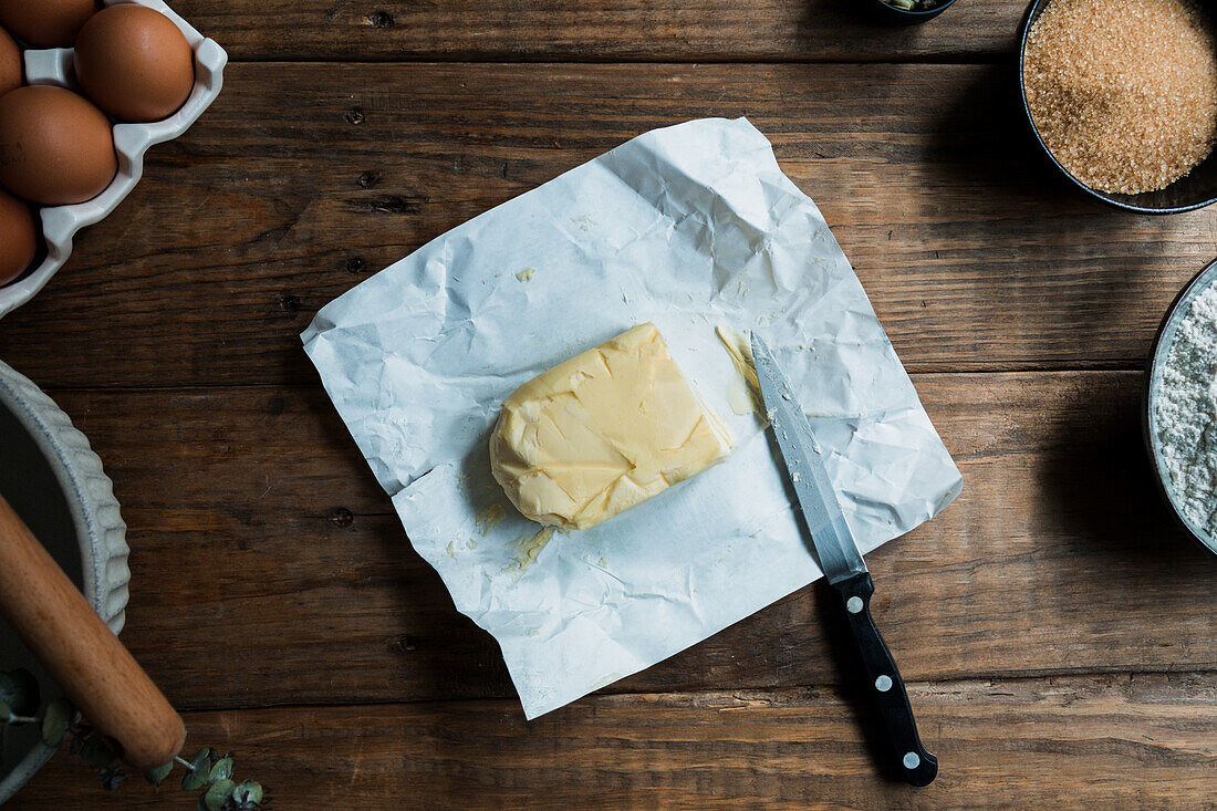 Messer bereit zum Schneiden eines Stücks Butter auf einem Gebäckstück auf einem Holztisch neben Eiern und braunem Zucker
