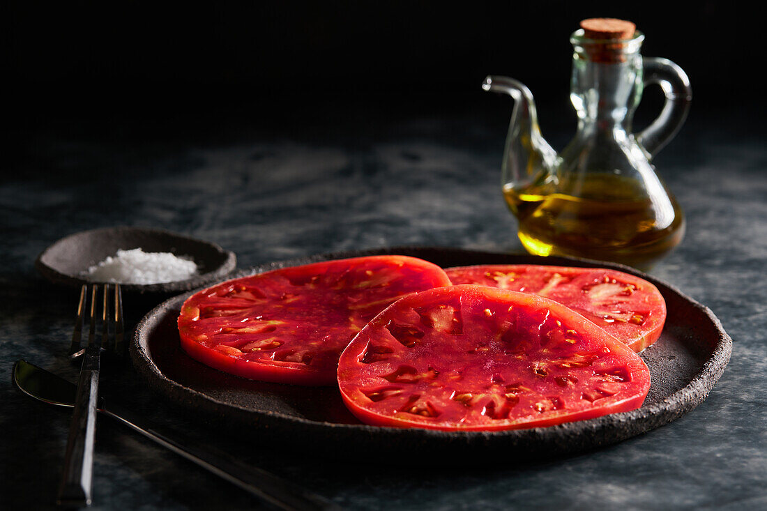 Delicious sliced tomatoes in cast iron plate near sea salt and jug of olive oil on concrete table