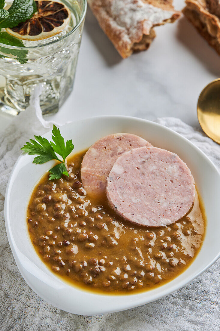 From above spoon placed near bowl of yummy lentil soup served with slices of sausage and leaf of parsley on marble table and gray napkin