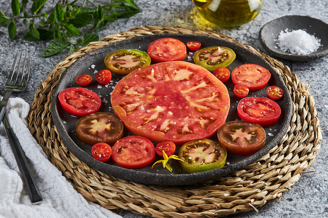 Delicious sliced tomatoes in cast iron plate placed on wicker napkin near plate with sea salt and jug of olive oil on concrete table