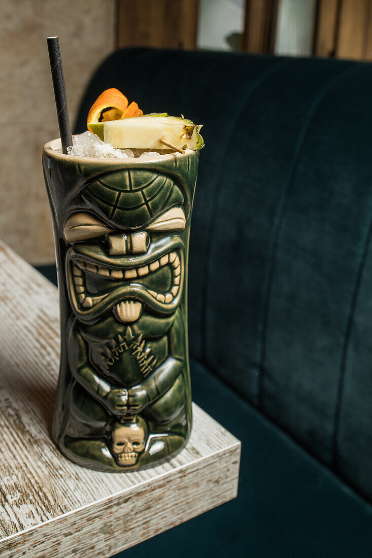 Large sculptural tiki cup filled with booze decorated with straw and fruits placed on green rug against wooden table