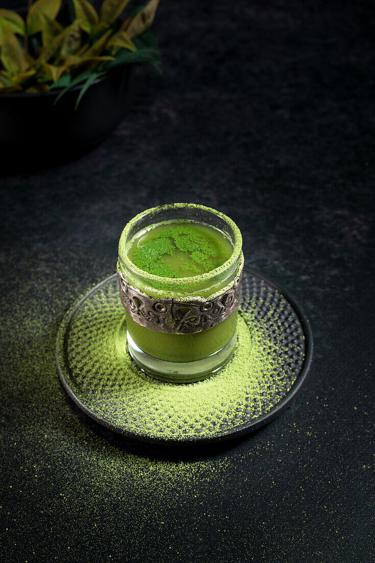 From above of healthy herbal green matcha tea served in glass cup with metal decoration on saucer sprinkled with powder on black table