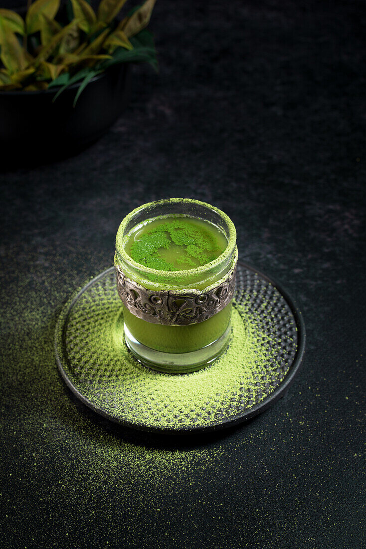 Von oben gesunder grüner Matcha-Kräutertee, serviert in einer Glastasse mit Metalldekoration auf einer mit Pulver bestreuten Untertasse auf einem schwarzen Tisch