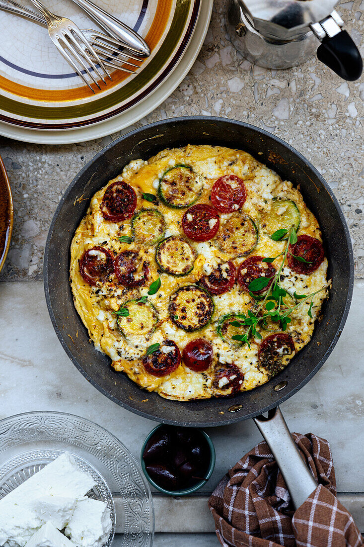 Top view of traditional Greek frittata with cherry tomatoes and feta cheese in frying pan with bread in light kitchen