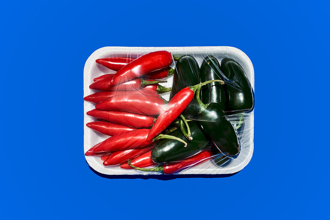 Top view of ripe chili and green spicy pepper in plastic wrap prepared for consumption on blue background