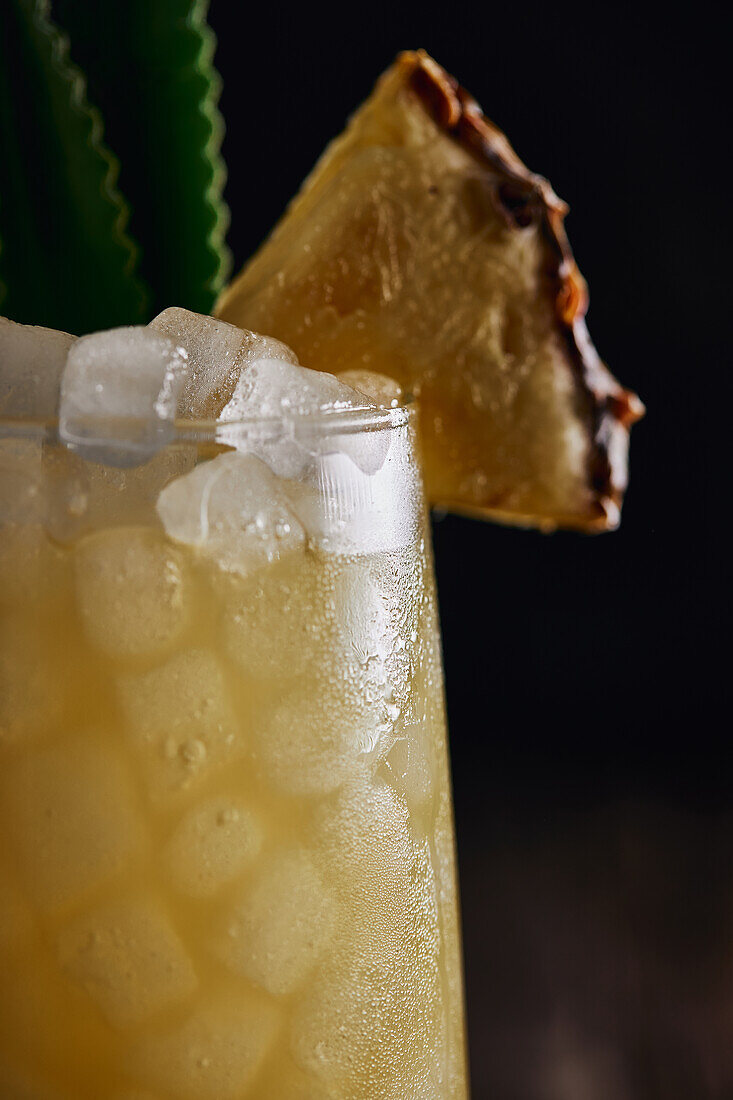 Holztisch mit gelbem Cocktailglas mit Eiswürfeln und erfrischendem Cocktail, garniert mit stacheligen Blättern und gestreiftem Stroh