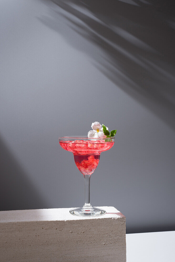 Crystal glass of pomegranate margarita cocktail served with flower blooms and leaf on concrete blocks in studio