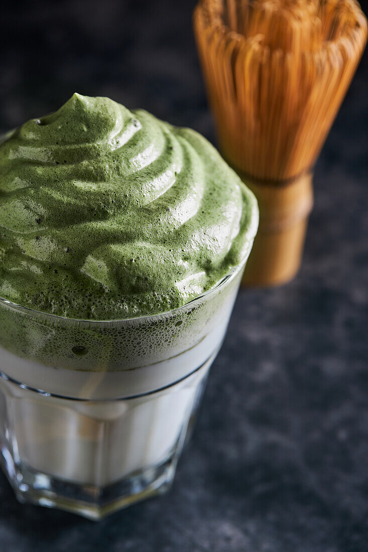 Glas frischer Dalgona-Kaffee mit Matcha und japanisches Teezeremonie-Zubehör, bestehend aus einem Bambusbesen namens Chasen, auf einem Holztisch