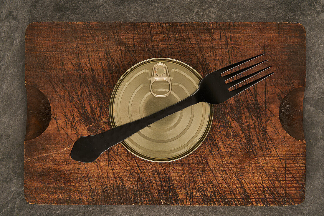 From above scratched chopping board with fork and sealed can with preserved food on rustic lumber table