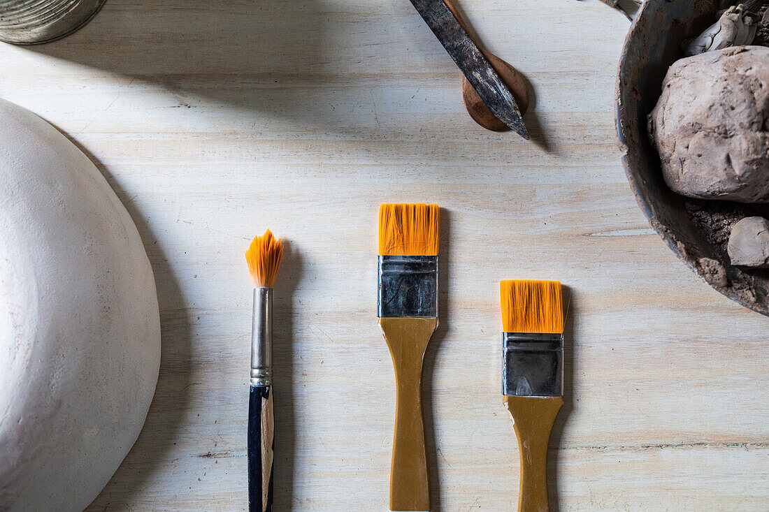 Top view set of pottery and painting brushes placed on wooden table with bowl of clay and ceramic pot in workshop