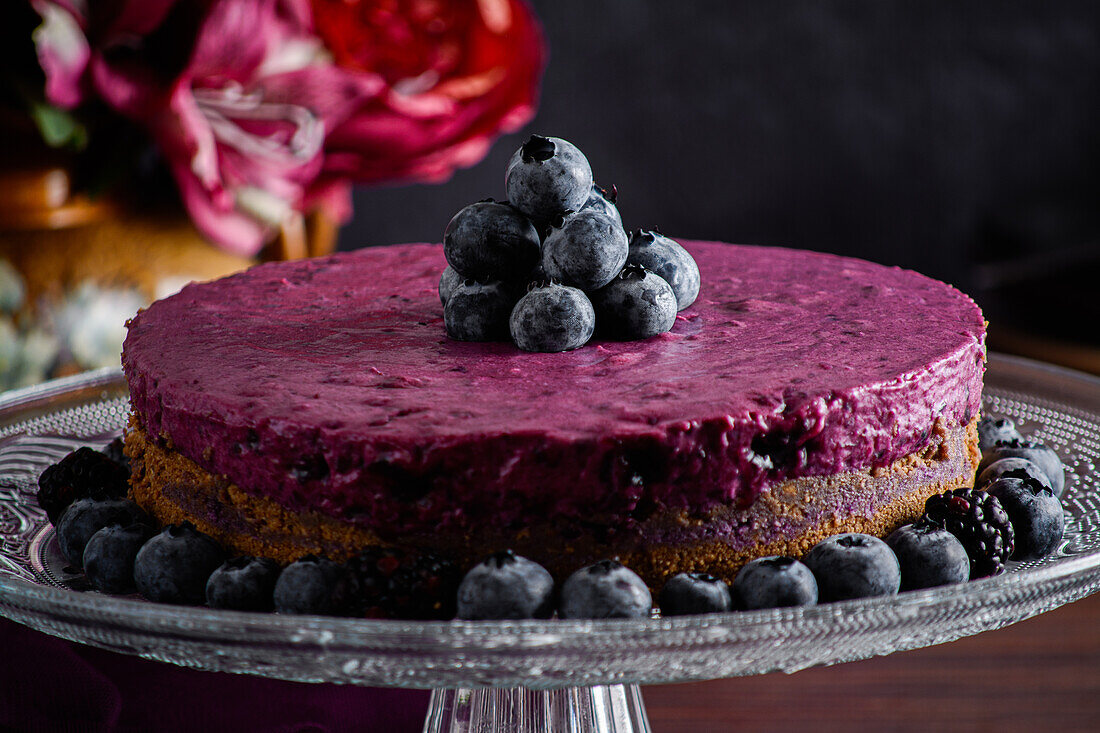 Delicious blueberry mousse cake with purple cream decorated with fresh berries served on glass stand on dark table with flowers