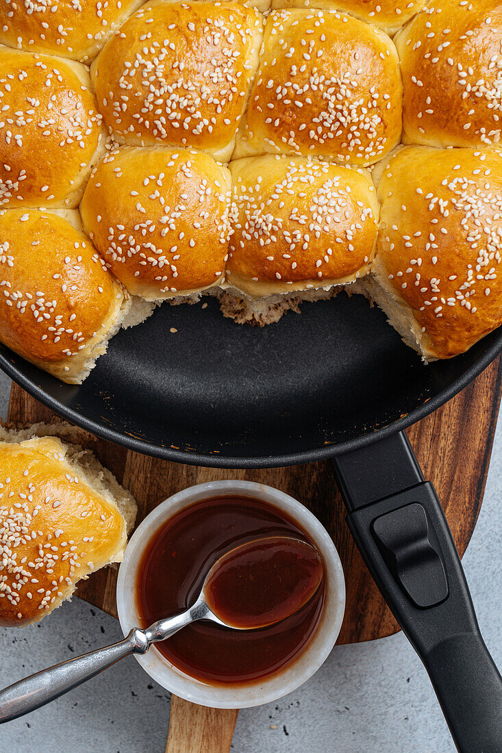 From above of tasty pull apart bread rolls with sesame seeds in frying pan served on wooden cutting board with sweet sauce