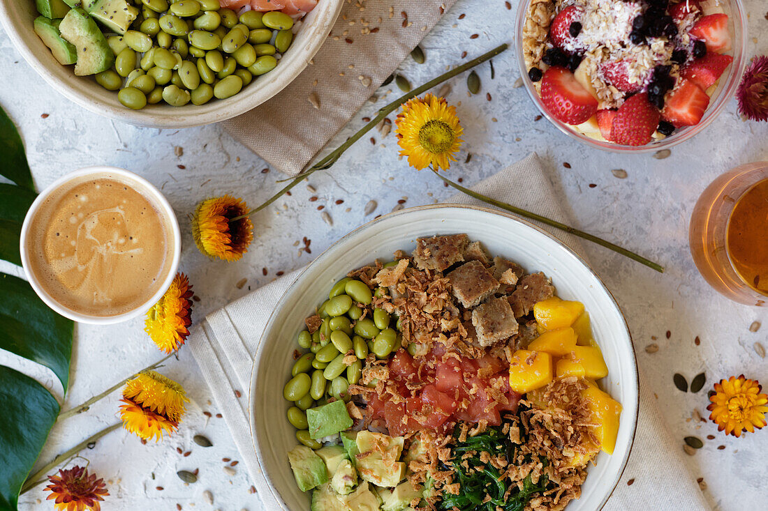 Top view appetizing assorted poke bowls with avocado toasts and glasses of juices served on table with berry dessert near scattered fresh flowers