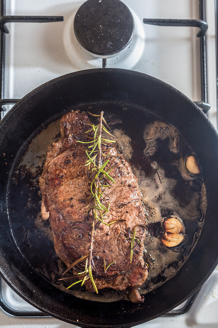From above of pan with appetizing meat piece with condiments and rosemary sprig in melted butter on gas stove