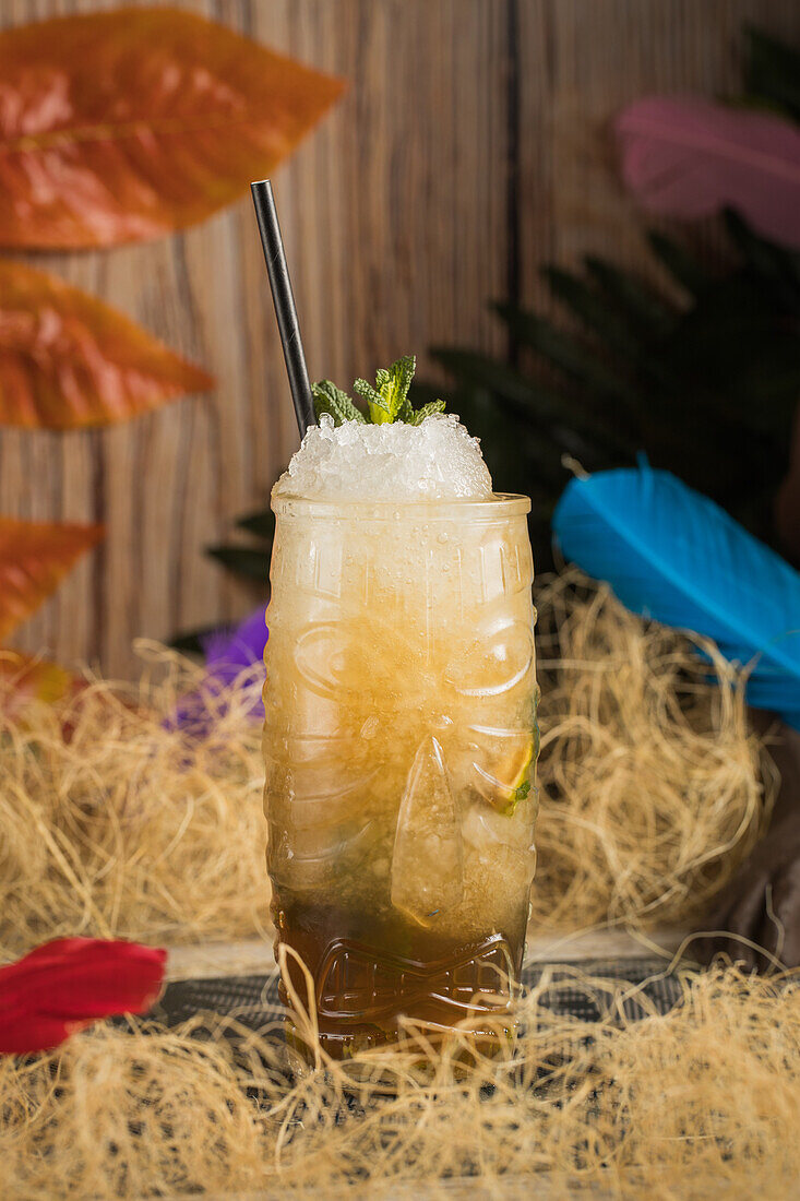 Tiki-Becher mit kaltem alkoholischem Getränk mit Strohhalm, serviert mit Eis und dekoriert mit frischen Kräutern, vor trockenem Gras auf unscharfem Hintergrund