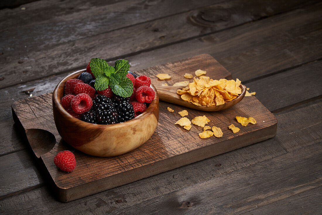 Spoon with cornflakes near bowl with heap of various ripe berries on wooden chopping board in kitchen