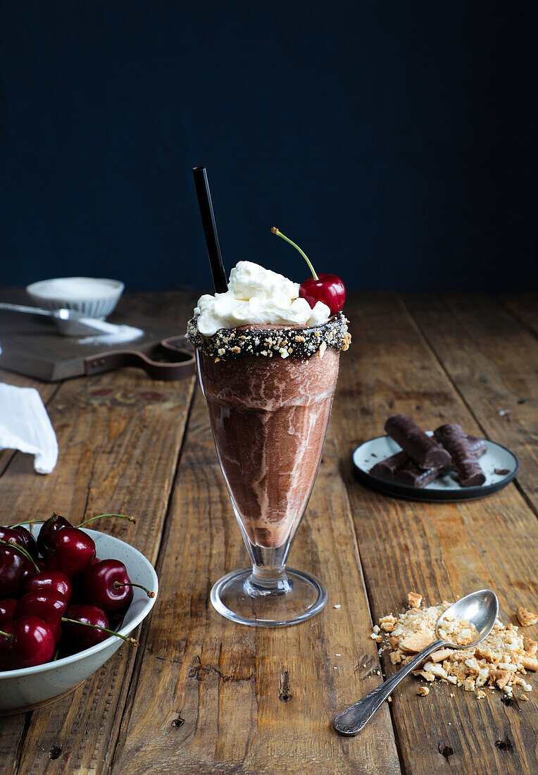 Cup of chocolate milkshake topped with cream and a cherry placed on wooden surface against a dark background