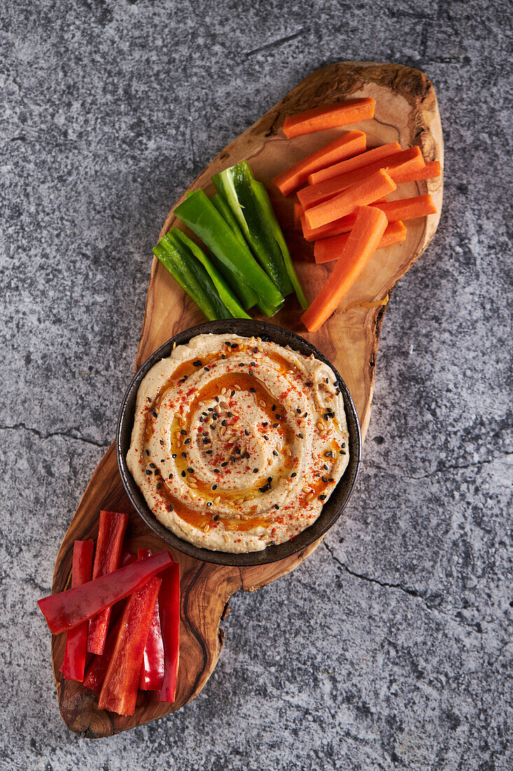 Draufsicht auf köstlichen Hummus und verschiedene Gemüsescheiben, die auf einem Schneidebrett auf einem grauen Marmortisch serviert werden