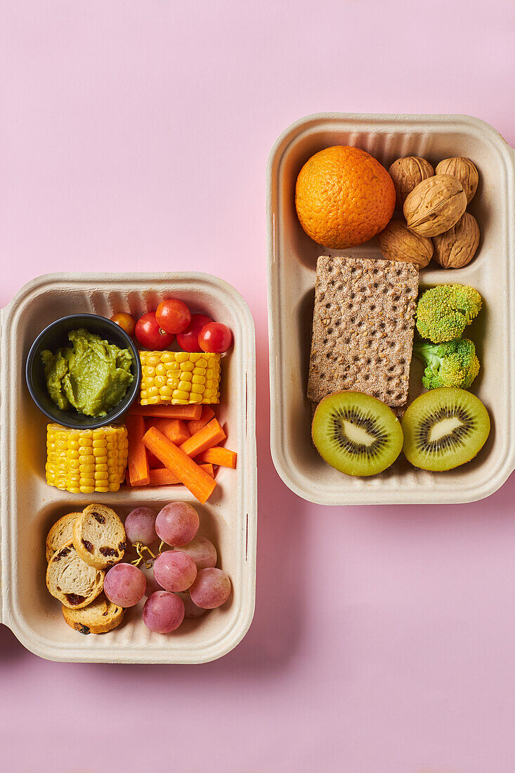 From above of lunch boxes with healthy food including crackers carrot sticks grapes cherry tomatoes with kiwi broccoli walnut and tangerine on pink background