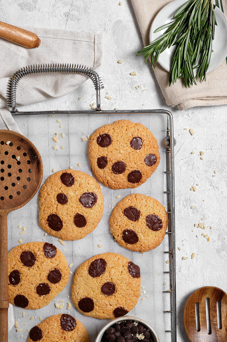 Top view of freshly baked sweet cookies with chocolate chips on metal grid placed on table with various kitchen tools and green rosemary branches