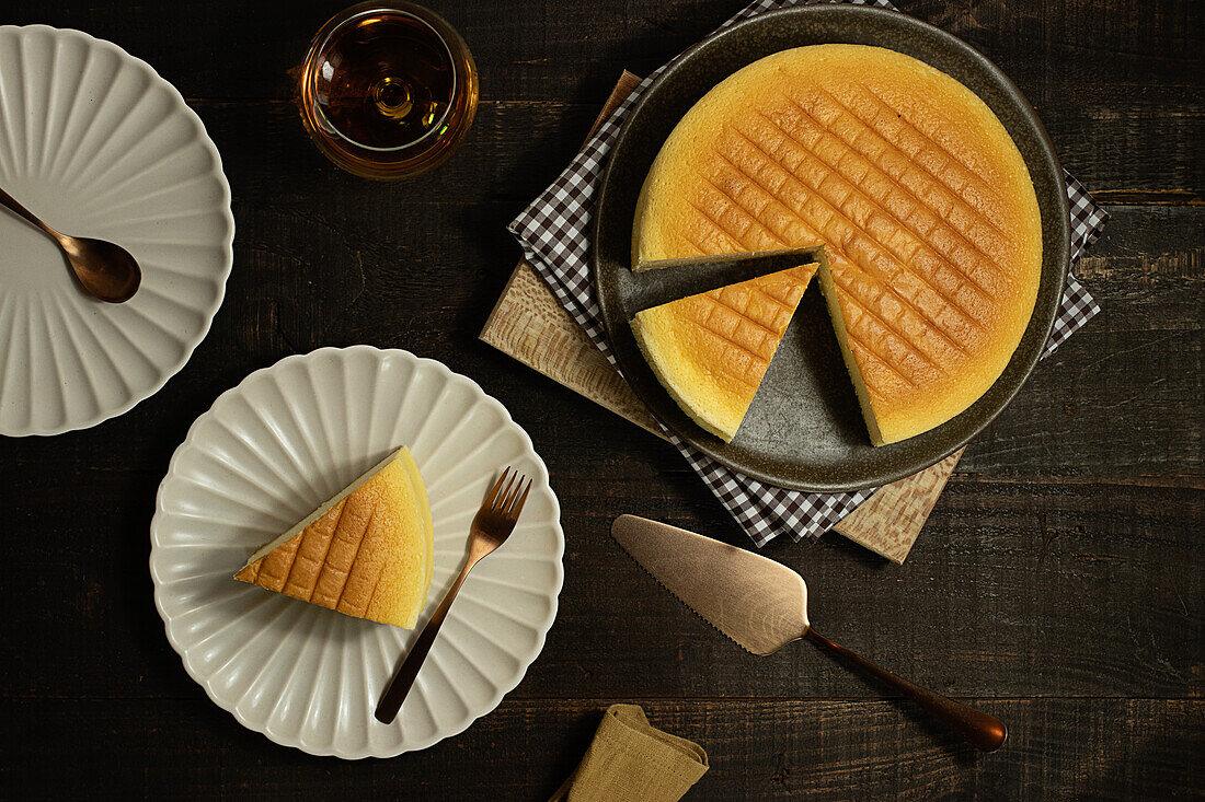 From above plates with tasty cotton cheesecake placed near glass of beverage on wooden table in dark room