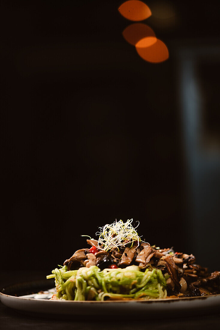 Niedriger Winkel eines leckeren veganen Gerichts mit Zucchini-Spaghetti und sautierten Pilzscheiben, bedeckt mit roten Beeren und Alfalfa-Sprossen auf dunklem Hintergrund